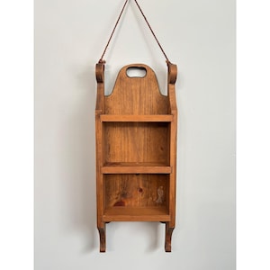 May include: A hanging wooden shelf with three tiers. The shelf is made of stained wood and has a handle at the top for hanging. The shelf is designed to be mounted on a wall and is a warm brown color.