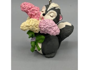May include: A skunk figurine holding a bouquet of flowers. The skunk is black and white with a pink nose and closed eyes. The flowers are pink, yellow, and purple, tied with a green and purple bow. A silver loop is at the top for hanging.