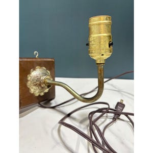 Peut inclure: Une applique murale composée d'une base carrée en bois foncé avec un petit crochet métallique sur le dessus. Un bras incurvé en laiton terni s'étend depuis une plaque métallique festonnée, menant à une douille cylindrique en laiton.