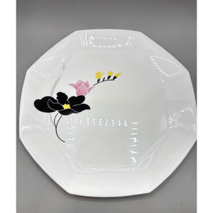 May include: A white octagonal plate featuring a floral design with a large black flower, a smaller pink flower, and yellow accents. The plate has a smooth, glossy finish and a distinct geometric shape with eight sides, providing a clean surface.