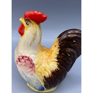 Puede incluir: Figura de gallo de cerámica con cresta y barbilla rojas, plumas de la cola marrones y cuerpo amarillo y blanco. El gallo tiene un diseño detallado y es probablemente un objeto decorativo.