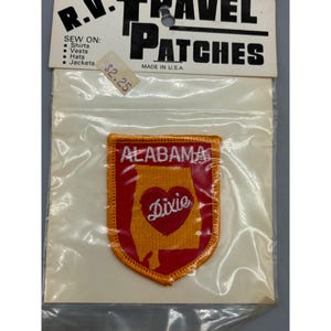 Puede incluir: Un parche bordado rojo y naranja que presenta el contorno del estado de Alabama con un corazón rojo en su interior que dice "Dixie". El parche está en una tarjeta blanca con el texto "R.V. TRAVEL PATCHES" y "MADE IN U.S.A."