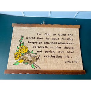May include: A rectangular wall hanging made of tan burlap fabric held by two horizontal wooden slats. The fabric features a printed illustration of a closed book with yellow roses and the text: For God so loved the world, that he gave his only begotten son, that whosoever believeth in him should not perish, but have everlasting life. John 3:16.