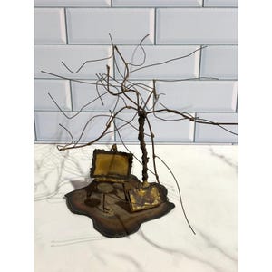 May include: A metal sculpture featuring a twisted wire tree structure standing on a dark, irregular base. Beside the tree is a small, rectangular gold-toned easel and a matching gold-toned stool, both crafted from metal with a weathered finish.