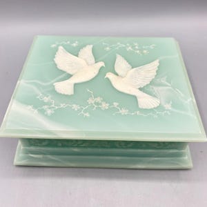 May include: A square, pale green jewellery box with a white marble effect. Two white doves are embossed on the lid, along with floral accents. The box has a rectangular base and a matching lid.