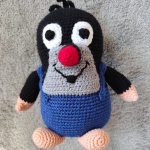 Può includere: Un giocattolo di peluche all'uncinetto fatto a mano di una talpa dei cartoni animati. La talpa ha una testa nera con grandi occhi bordati di bianco, un naso rosso e una faccia grigia. Indossa un completo blu con braccia nere e mani e piedi color pesca.