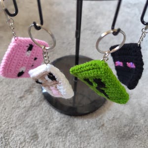 Peut inclure: Une collection de porte-clés crochetés à la main. Les porte-clés présentent divers motifs, dont un cochon rose, une vache blanche et marron, un creeper vert et un enderman noir. Chaque porte-clés est attaché à un porte-clés argenté.