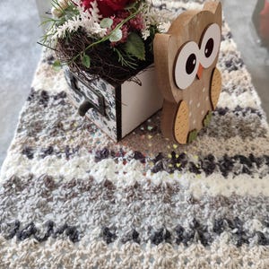 Op de afbeelding: Een gehaakte tafelloper in tinten wit, bruin en grijs met een houten uilbeeldje en een rode roos in een decoratieve lade.