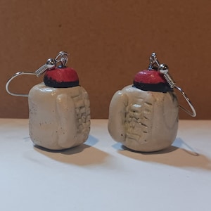 Puede incluir: Un par de pendientes hechos a mano con pequeñas mochilas de cerámica blanca texturizada con detalles rojos y negros. Las mochilas tienen un cierre de gancho plateado.