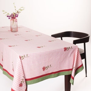 May include: A pink and white gingham tablecloth with a green and red border. The tablecloth has a floral pattern with red flowers and green stems. A vase with purple flowers sits on the tablecloth.