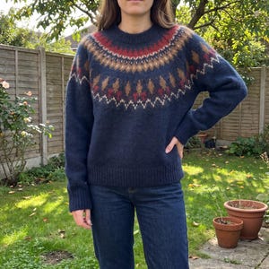 May include: A navy blue long-sleeve sweater featuring a round neckline and a decorative yoke pattern in shades of red, tan, and cream. The knit garment has ribbed cuffs and a ribbed hem, providing a textured finish to the overall design.