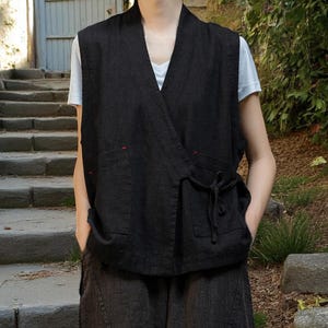 May include: Black wrap vest with two front pockets and a tie closure. The vest has red stitching accents and is worn over a white t-shirt. The vest is paired with matching black trousers.