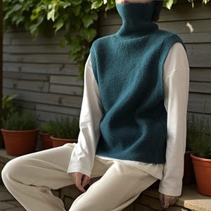 May include: A teal turtleneck sweater vest worn over a white long-sleeved shirt. The person is wearing cream corduroy trousers. The background features a wooden fence, greenery, and terracotta plant pots. The style is casual and comfortable.