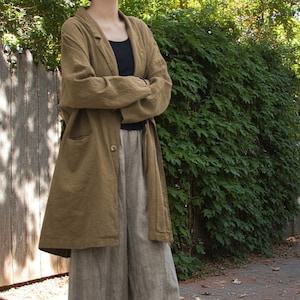 Peut inclure: Un long manteau en lin vert olive avec une fermeture &agrave; un bouton, port&eacute; sur un haut noir et un pantalon en lin gris clair. La tenue est ample, sugg&eacute;rant une esth&eacute;tique d&eacute;contract&eacute;e et confortable. L'arri&egrave;re-plan pr&eacute;sente une cl&ocirc;ture en bois et une v&eacute;g&eacute;tation luxuriante.