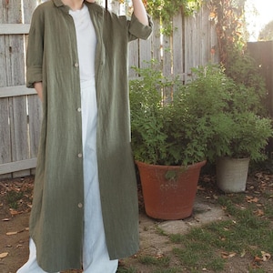 Puede incluir: Abrigo largo de lino verde oliva con botones. El abrigo largo se lleva sobre una camiseta blanca y pantalones blancos. El conjunto está diseñado al aire libre con una valla de madera y plantas en macetas en el fondo.