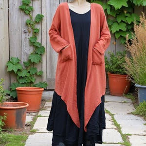May include: A long, flowing, terracotta-coloured cardigan with large pockets is worn over a black dress. The cardigan is made of a textured fabric. The outfit is worn outdoors in a garden setting with a wooden fence and potted plants.