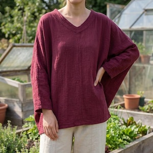 May include: A deep burgundy long-sleeve top with a V-neckline and a textured, crinkled fabric finish. The garment features a loose, relaxed fit with dropped shoulders and wide sleeves, paired with light beige trousers that have a central seam.