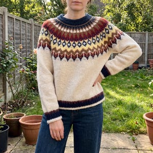 May include: A cream-coloured long-sleeve jumper featuring a round neckline and a yoke decorated with a geometric pattern in shades of dark blue, yellow, brown, and white. The cuffs and hem are finished with a dark blue ribbed knit trim.
