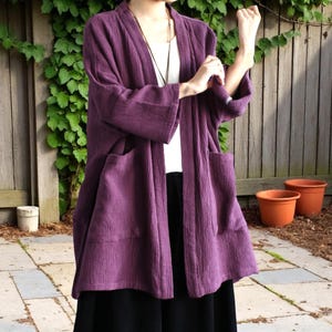 May include: A long, open-front purple jacket with large pockets. The jacket is made of a textured fabric and is worn over a white top and black skirt. The background includes a wooden fence, greenery, and terracotta pots.