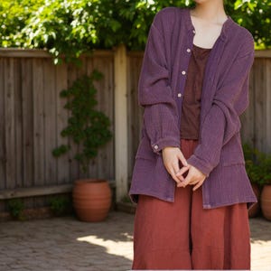 May include: An outfit comprising a long-sleeved, button-down, purple jacket, a brown top, and rust-coloured trousers. The jacket has a textured finish. The ensemble is worn outdoors, with a wooden fence and foliage in the background.