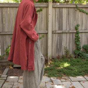Peut inclure: Un long manteau en lin couleur rouille avec une fermeture &agrave; bouton unique. Le manteau est associ&eacute; &agrave; un pantalon en lin gris clair ample. La tenue est pr&eacute;sent&eacute;e sur fond de cl&ocirc;ture en bois et de verdure, sugg&eacute;rant un cadre ext&eacute;rieur d&eacute;contract&eacute;.