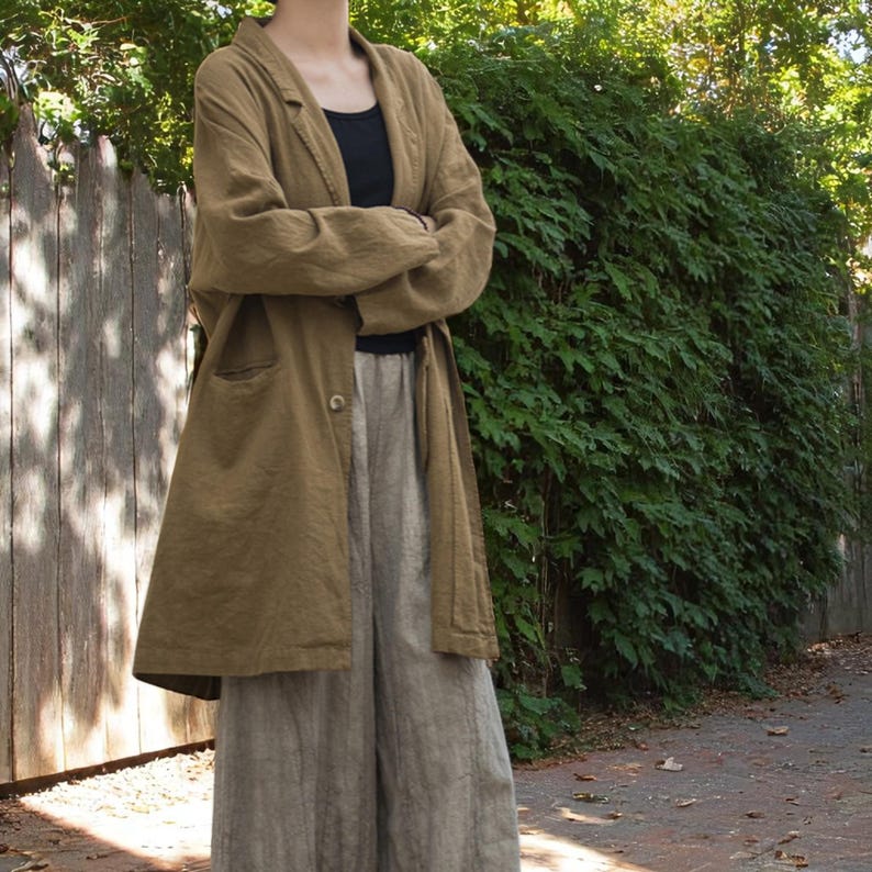 Peut inclure: Un long manteau en lin vert olive avec une fermeture &agrave; un bouton, port&eacute; sur un haut noir et un pantalon en lin gris clair. La tenue est ample, sugg&eacute;rant une esth&eacute;tique d&eacute;contract&eacute;e et confortable. L'arri&egrave;re-plan pr&eacute;sente une cl&ocirc;ture en bois et une v&eacute;g&eacute;tation luxuriante.