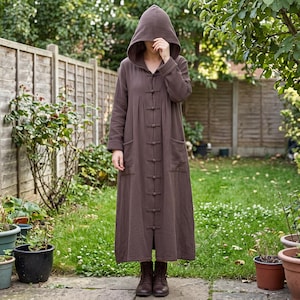May include: A long-sleeved, floor-length brown hooded coat featuring a front button closure with traditional toggle-style fasteners, two large patch pockets on the front, and a textured fabric finish, worn with dark brown ankle boots.
