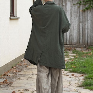 Peut inclure: Un manteau en lin vert fonc&eacute; avec un col et des manches longues. Le manteau est port&eacute; sur un pantalon en lin beige clair. La personne se tient sur un chemin pav&eacute; devant une cl&ocirc;ture en bois et un mur blanc.