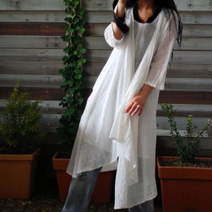 May include: A white, flowing tunic dress with long sleeves and a draped front. The dress is paired with blue jeans. The background features a wooden wall and potted plants, creating a casual, bohemian aesthetic.