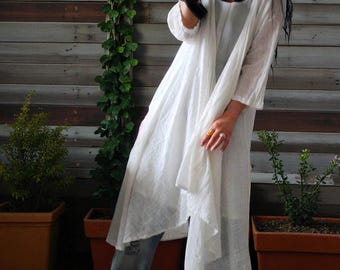 May include: A white, flowing tunic dress with long sleeves and a draped front. The dress is paired with blue jeans. The background features a wooden fence and potted plants, creating a casual, bohemian aesthetic.