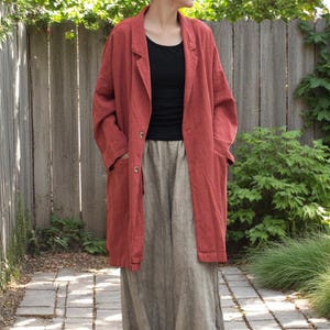 Peut inclure: Un long blazer en lin couleur rouille, avec deux boutons et des poches. Le blazer est port&eacute; sur un haut noir et une longue jupe de couleur claire. La tenue est d&eacute;contract&eacute;e et confortable, adapt&eacute;e au quotidien. L'arri&egrave;re-plan comprend une cl&ocirc;ture en bois et de la verdure.