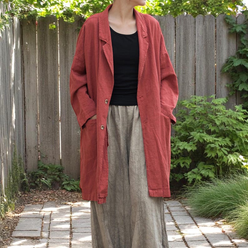 Peut inclure: Un long blazer en lin couleur rouille, avec deux boutons et des poches. Le blazer est port&eacute; sur un haut noir et une longue jupe de couleur claire. La tenue est d&eacute;contract&eacute;e et confortable, adapt&eacute;e au quotidien. L'arri&egrave;re-plan comprend une cl&ocirc;ture en bois et de la verdure.