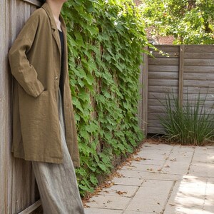 Peut inclure: Un long manteau en lin vert olive avec un seul bouton et deux poches avant, associ&eacute; &agrave; un pantalon en lin gris clair. En arri&egrave;re-plan, une cl&ocirc;ture en bois et un mur recouvert de vignes vertes. La longueur du manteau est d'environ 110 cm.