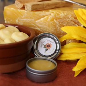 Puede incluir: Una naturaleza muerta con un gran girasol amarillo, un trozo de cera de abeja amarilla y un pequeño cuenco marrón lleno de objetos redondos de color amarillo pálido. También hay dos pequeñas latas de metal de bálsamo labial, una abierta, con la etiqueta "Sunflower Cupboard".