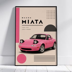 Op de afbeelding: Een ingelijste kunstprint met een roze Mazda Miata auto. De poster heeft een beige achtergrond met geometrische vormen en de tekst "MAZDA MIATA 1989-1997 LESS IS MORE". De auto heeft pop-up koplampen. De lijst is zwart.