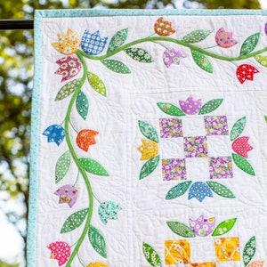 May include: A white quilt with a floral design. The quilt features a vine with green leaves and colorful tulips. The tulips are made from different patterned fabrics. The quilt is edged with a light blue trim.