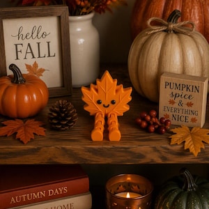 May include: A bright orange maple leaf figurine with a smiling face and dangling legs, positioned centrally. The leaf has black eyes and a small mouth, with a textured surface. It stands upright on a wooden surface among various autumn decorations.