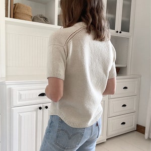 以下が含まれることがあります： クリーム色のニットトップス、半袖、装飾的なネックライン。明るいブルーデニムのジーンズと組み合わせています。画像は屋内撮影で、背景には白いキャビネットがあります。