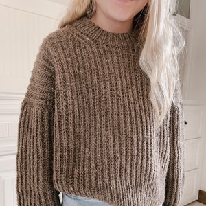 以下が含まれることがあります： クルーネックと長袖の茶色のリブ編みセーター。セーターはゆったりとしたフィット感で、ライトブルーのジーンズと組み合わせています。ニットウェアは、柔らかく質感のある糸で作られているようです。