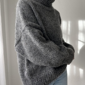 Puede incluir: Un su&eacute;ter gris de cuello alto con una textura peluda. El su&eacute;ter est&aacute; hecho de un material suave y c&aacute;lido y tiene un corte holgado.