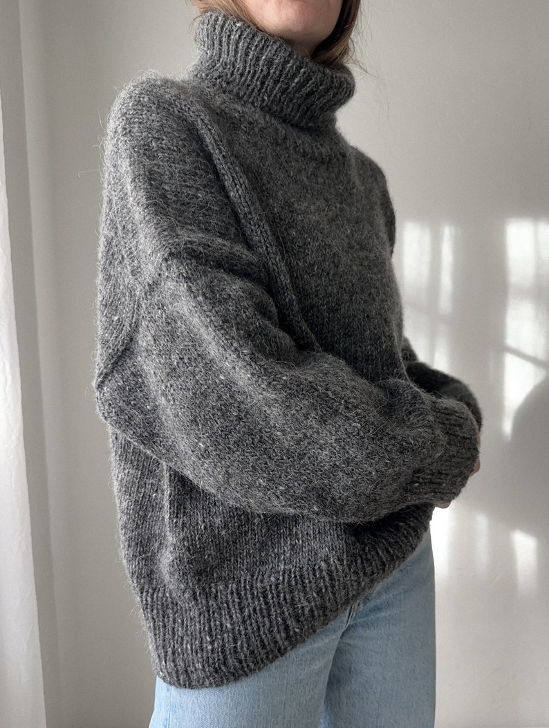 Puede incluir: Un su&eacute;ter gris de cuello alto con una textura peluda. El su&eacute;ter est&aacute; hecho de un material suave y c&aacute;lido y tiene un corte holgado.