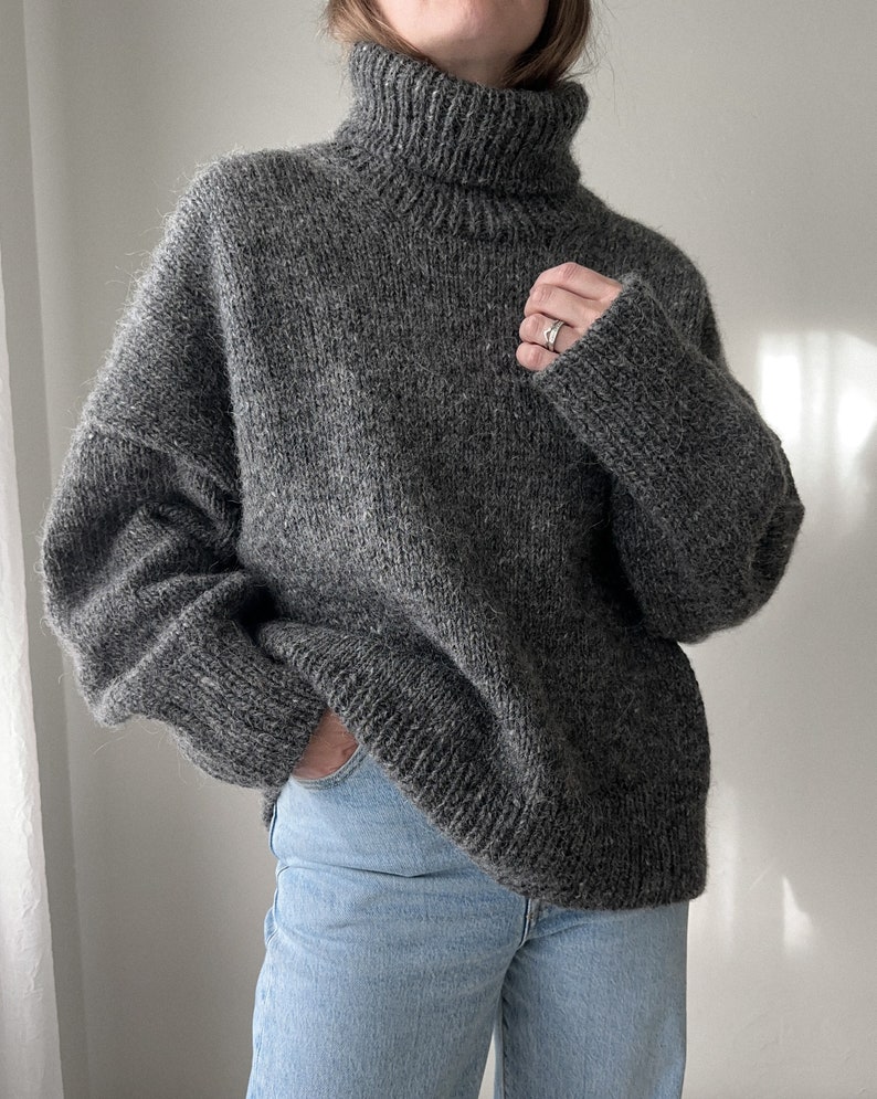Puede incluir: Un su&eacute;ter gris de cuello alto de gran tama&ntilde;o con un corte holgado. El su&eacute;ter est&aacute; hecho de un material suave y peludo.