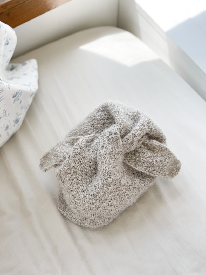 May include: A light gray knitted hat with a textured appearance, resting on a white surface. The hat is folded and knotted, with a soft, cozy aesthetic. The image is well-lit, highlighting the hat's details.