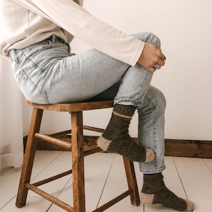 以下が含まれることがあります： 白い長袖シャツと薄いブルーのジーンズを着用した人が、木製の椅子に座っています。茶色とベージュのストライプの靴下を履いています。