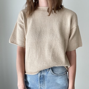 May include: A beige short-sleeved knitted top with a round neckline. The top is made of a soft, textured fabric and is a relaxed fit.