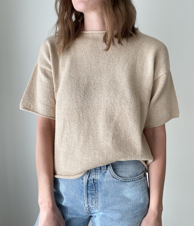 May include: A beige short-sleeved knitted top with a round neckline. The top is made of a soft, textured fabric and is a relaxed fit.