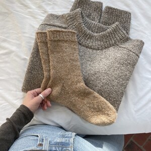 K&ouml;nnte beinhalten: Ein Paar braune Stricksocken und ein grauer Strickpullover. Die Socken werden in einer Hand gehalten, die ein braunes Hemd und eine blaue Jeans tr&auml;gt.