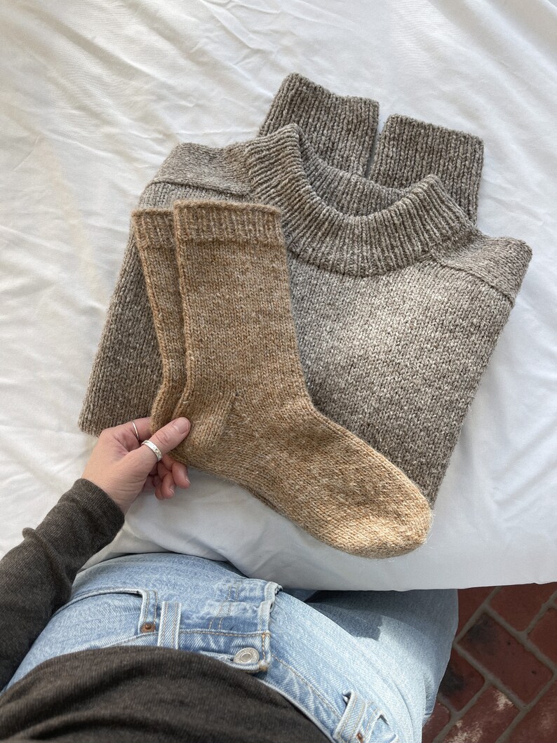 K&ouml;nnte beinhalten: Ein Paar braune Stricksocken und ein grauer Strickpullover. Die Socken werden in einer Hand gehalten, die ein braunes Hemd und eine blaue Jeans tr&auml;gt.