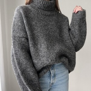 Puede incluir: Un su&eacute;ter gris de cuello alto con un corte holgado y mangas largas. El su&eacute;ter est&aacute; hecho de un material suave y peludo.