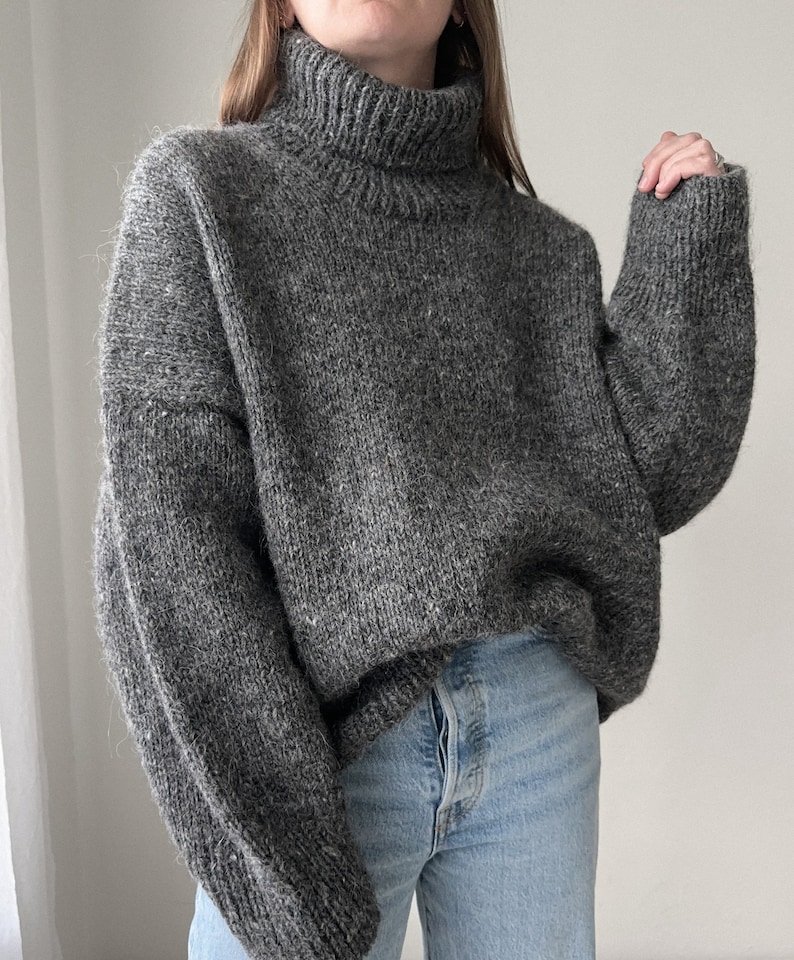Puede incluir: Un su&eacute;ter gris de cuello alto con un corte holgado y mangas largas. El su&eacute;ter est&aacute; hecho de un material suave y peludo.